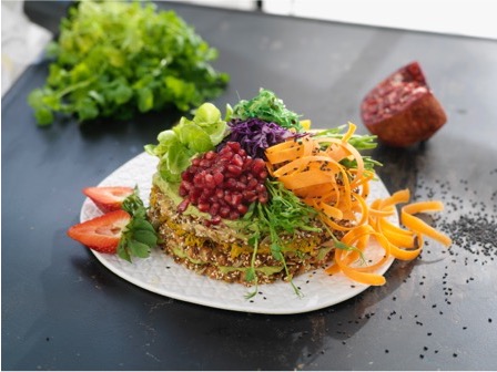 En inbjudande vegansk smörgåstårta med flera lager av krämiga röror och mjukt bröd, täckt med ett jämnt lager växtbaserad färskost. Ovansidan är dekorerad med färska grönsaker som skivade gurkor, körsbärstomater, avokado och örter, vilket ger en färgstark och fräsch presentation.