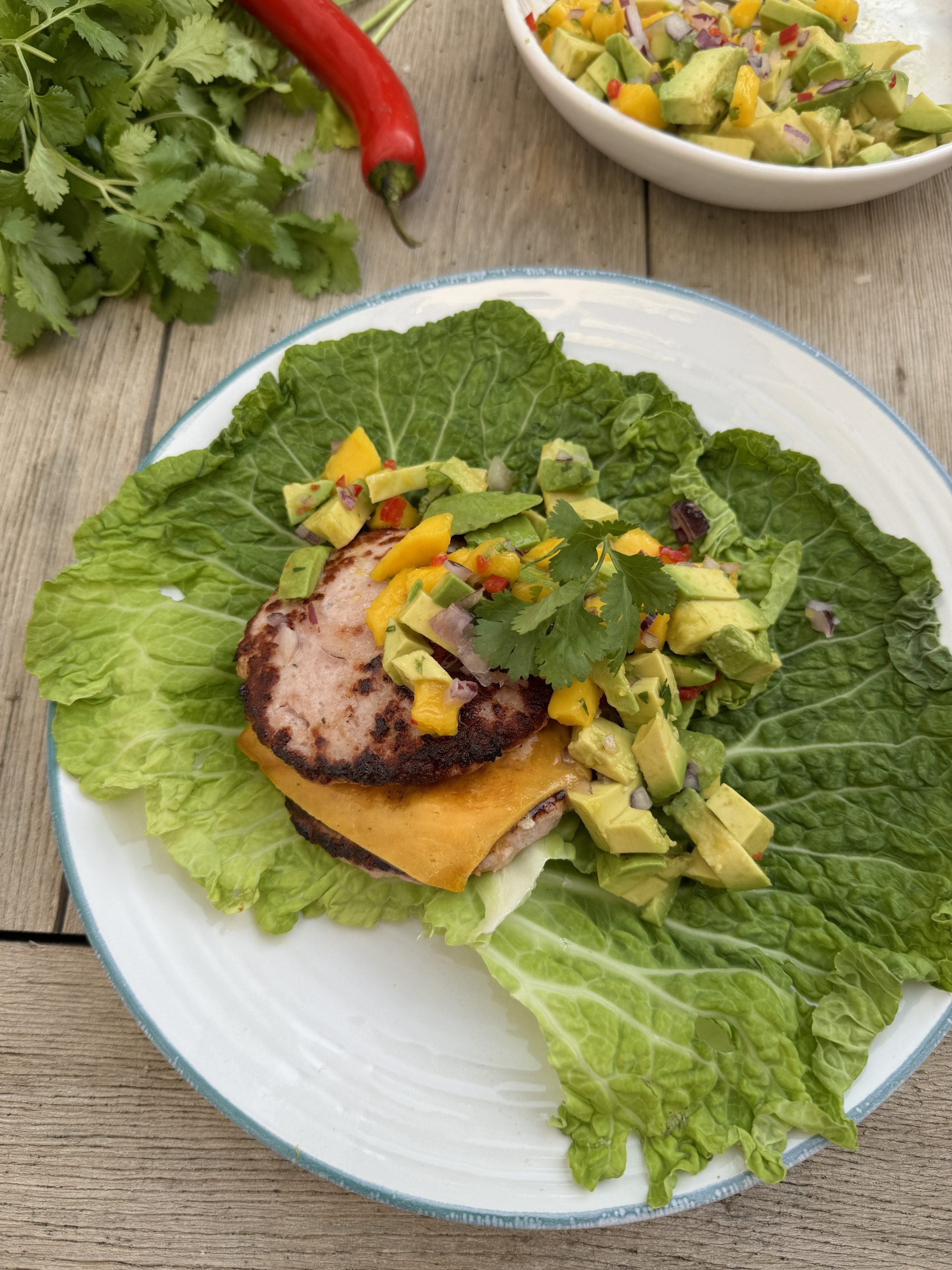 En mexikansk hamburgare utan bröd, serverad på krispiga salladsblad ovanpå en vit tallrik. Hamburgaren är toppad med smält ost, skivor av jalapeño, avokado och färgstark salsa, vilket ger en fräsch och färgstark presentation.